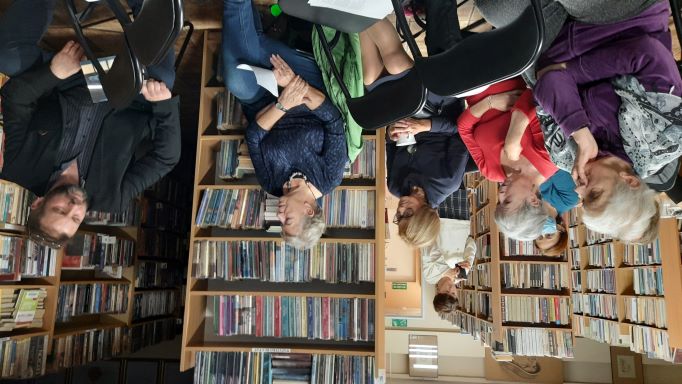 Zdjęcie przedstawia siedzących ludzi w sali bibliotecznej, mężczyznę i kilka kobiet.