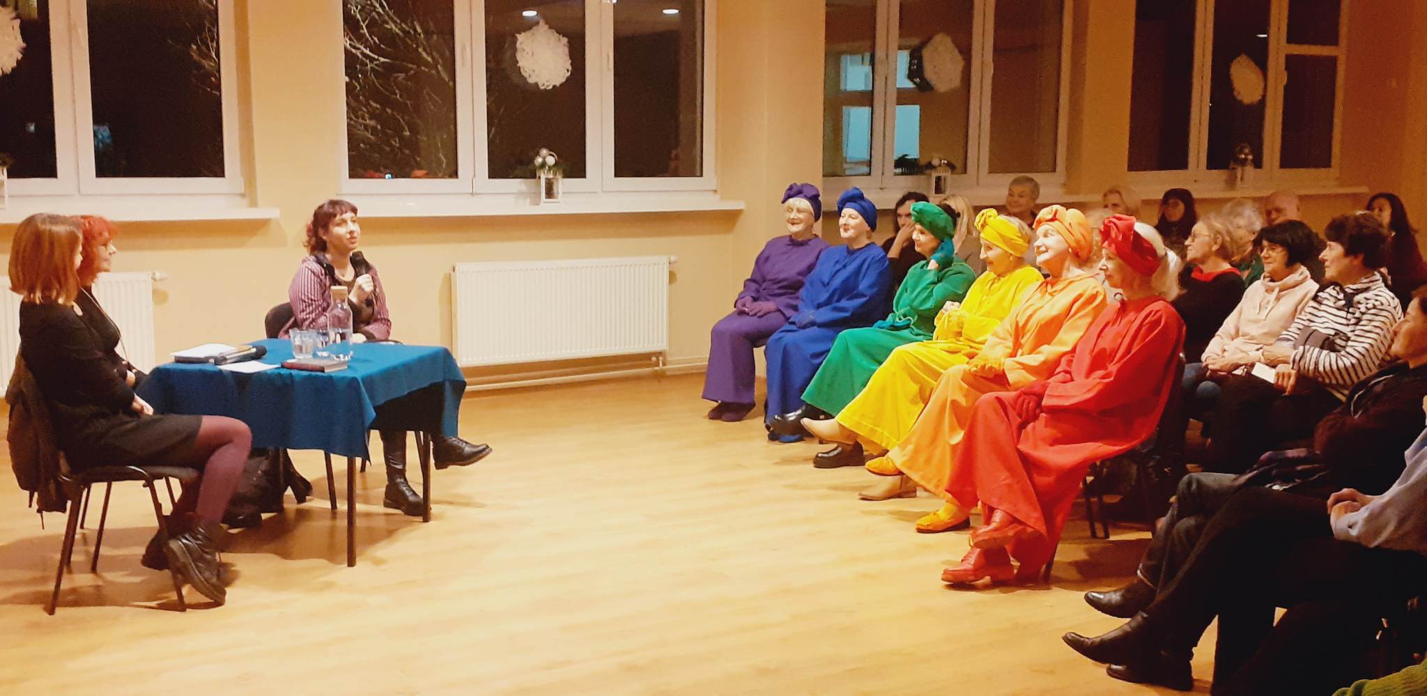 Zdjęcie przedstawia siedzących ludzi w sali, przy stoliku siedzą trzy kobiety, na przeciwko nich kobiety ubrane w kolory tęczy